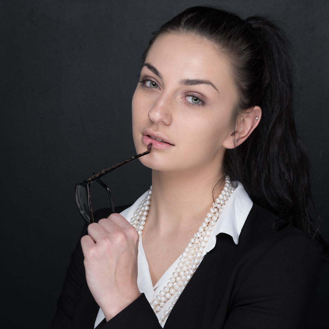 Person in a black blazer and white shirt holding glasses to their lips, with a thoughtful expression against a dark background.