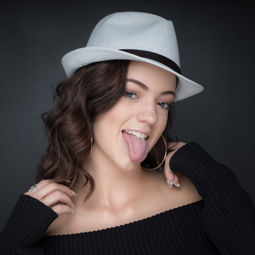 Person wearing a white fedora and black sweater, making a playful gesture with their tongue out, against a dark background.