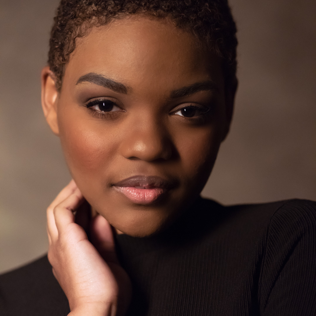 Person with short hair and subtle makeup, wearing a black top, poses against a neutral background while touching their neck.