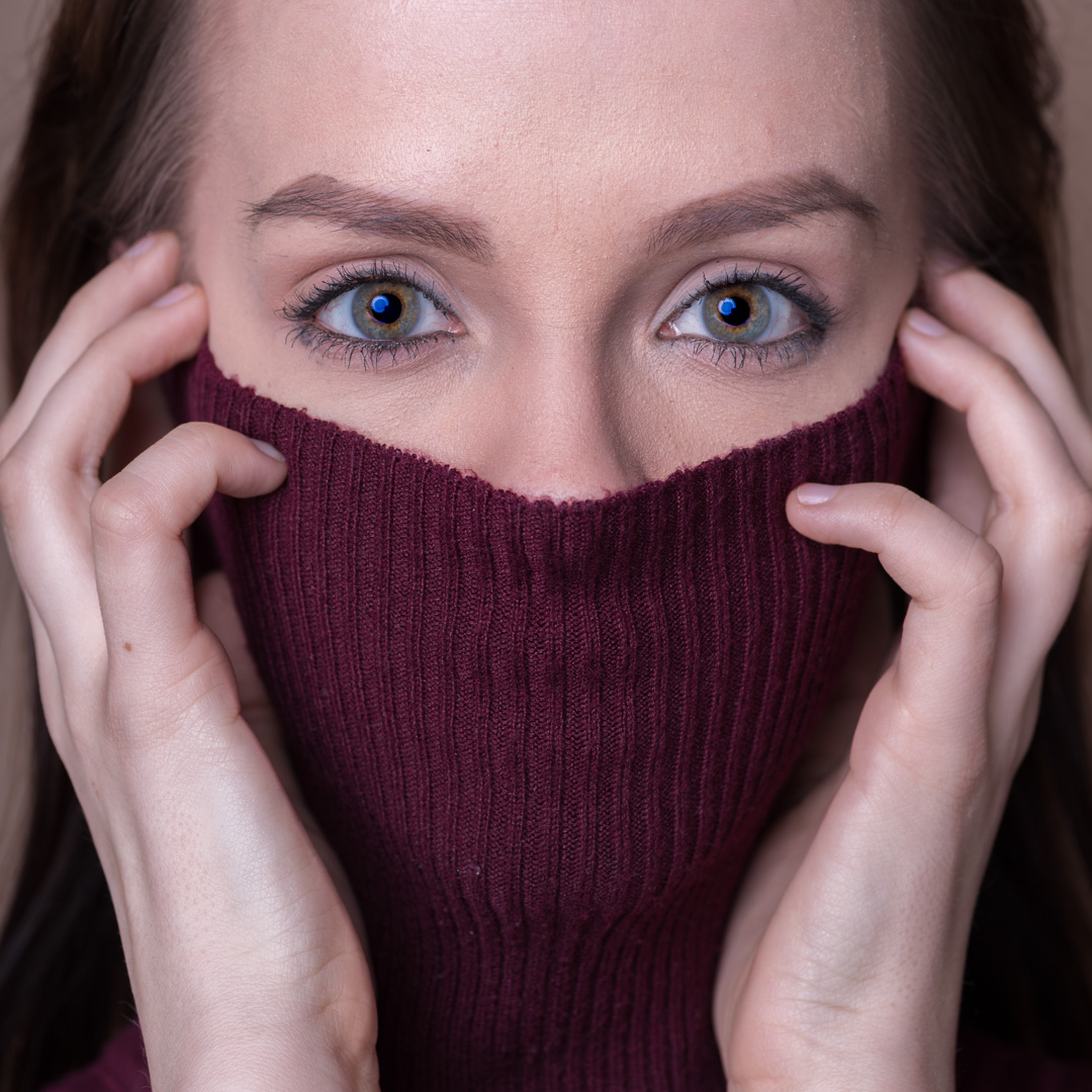 Person with blue eyes, partially covering their face with a burgundy sweater, holding it with both hands.