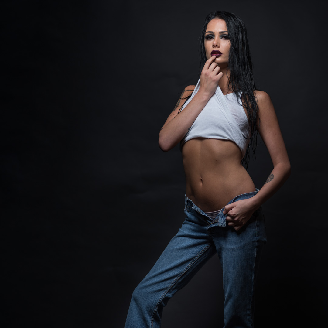 Person in jeans and a white crop top poses against a dark background, with one hand on their lips and the other adjusting the jeans.