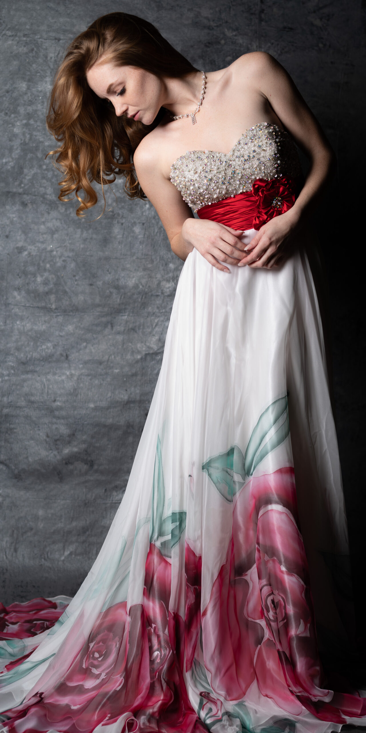 Woman in strapless gown with beaded bodice, red sash, and floral skirt standing against a textured gray background.