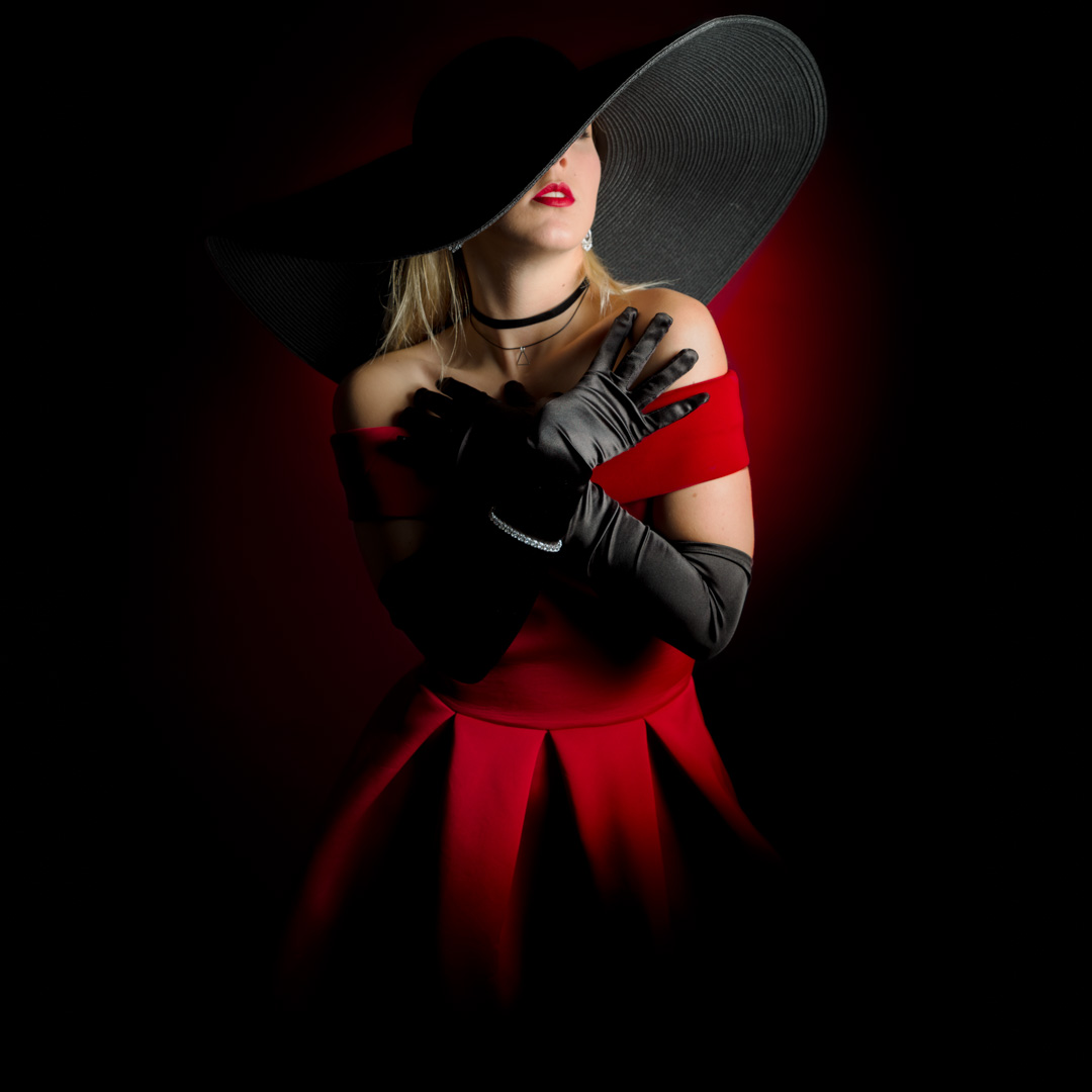 Person in a red dress and large black hat, wearing long black gloves, stands against a dark background with red lighting.