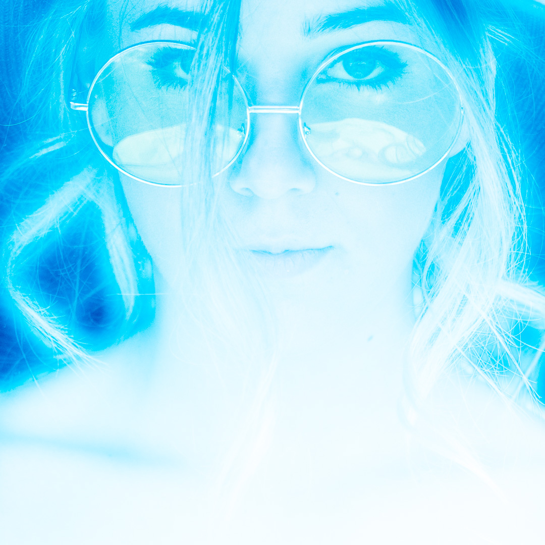 Close-up of a person wearing round glasses, with a soft blue filter and ethereal lighting highlighting facial features.