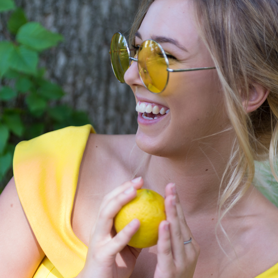 A woman with sunglasses and a yellow dress smiles while holding a lemon. She is outdoors near a tree.