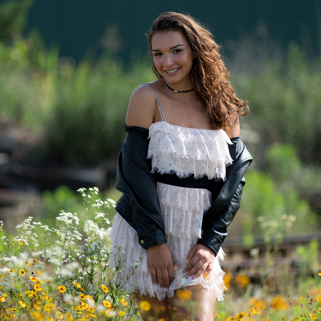 A person with long hair wearing a light lace dress and black leather jacket stands in a field of wildflowers, smiling at the camera.