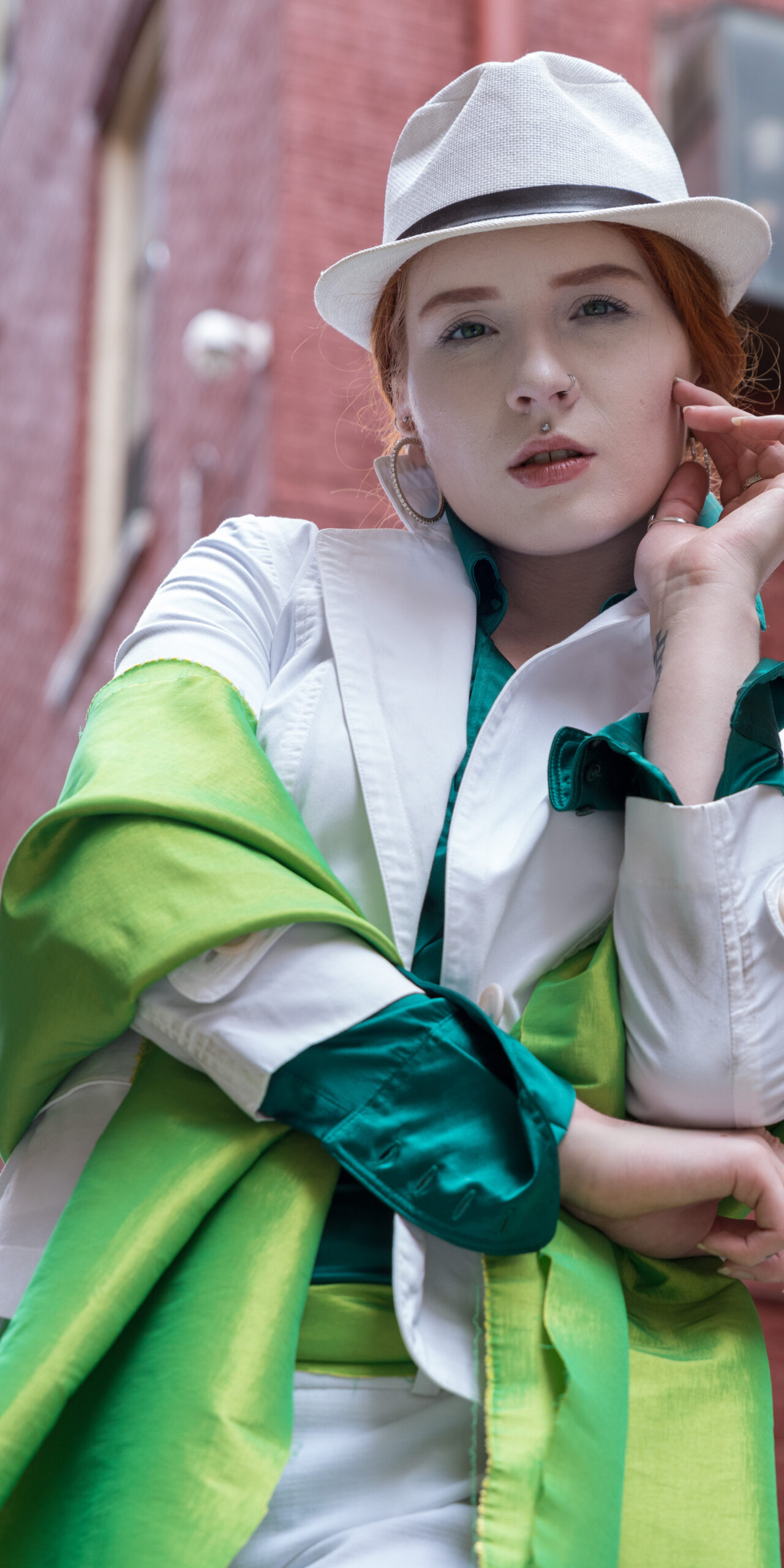 Person wearing a white hat and coat with a green shirt and scarf, standing in an alley with brick walls.