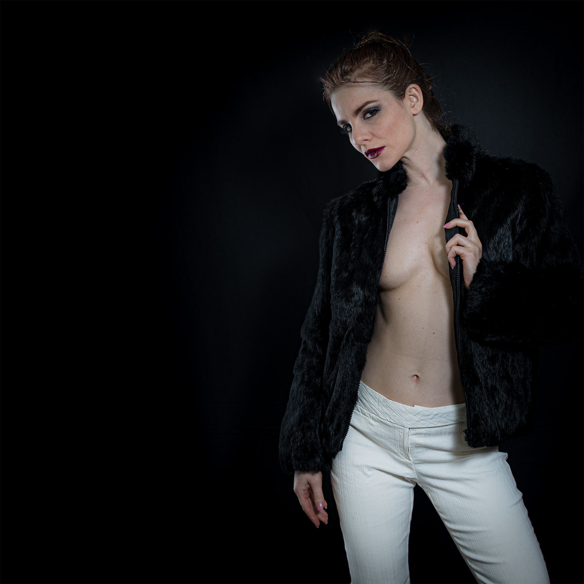 Person in a black fur jacket and white pants poses against a dark background.