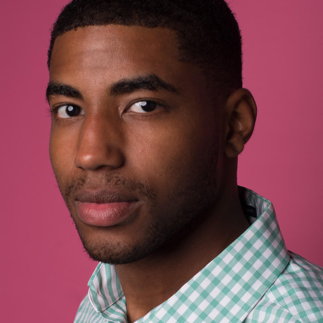 Person wearing a green checkered shirt, looking at the camera in front of a pink background.