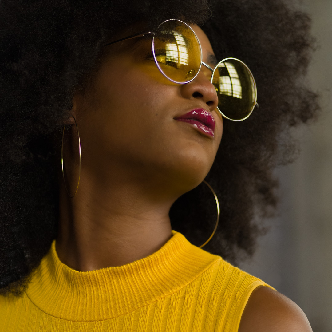 Person with large curly hair wearing yellow sunglasses, hoop earrings, and a yellow top, looking to the side.