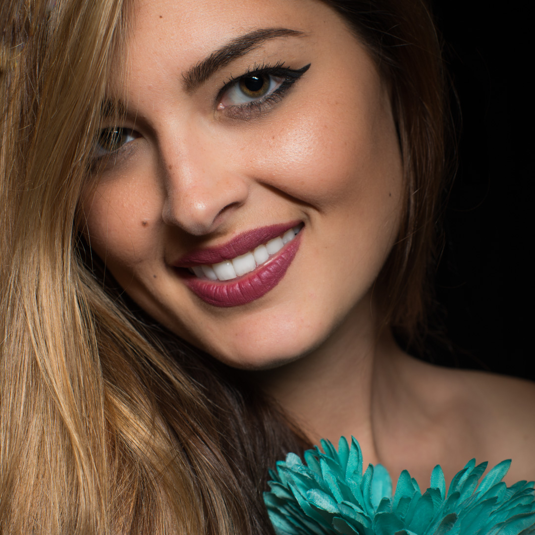 A woman with long hair smiling, wearing dark eyeliner and purple lipstick, holds a teal flower against a black background.