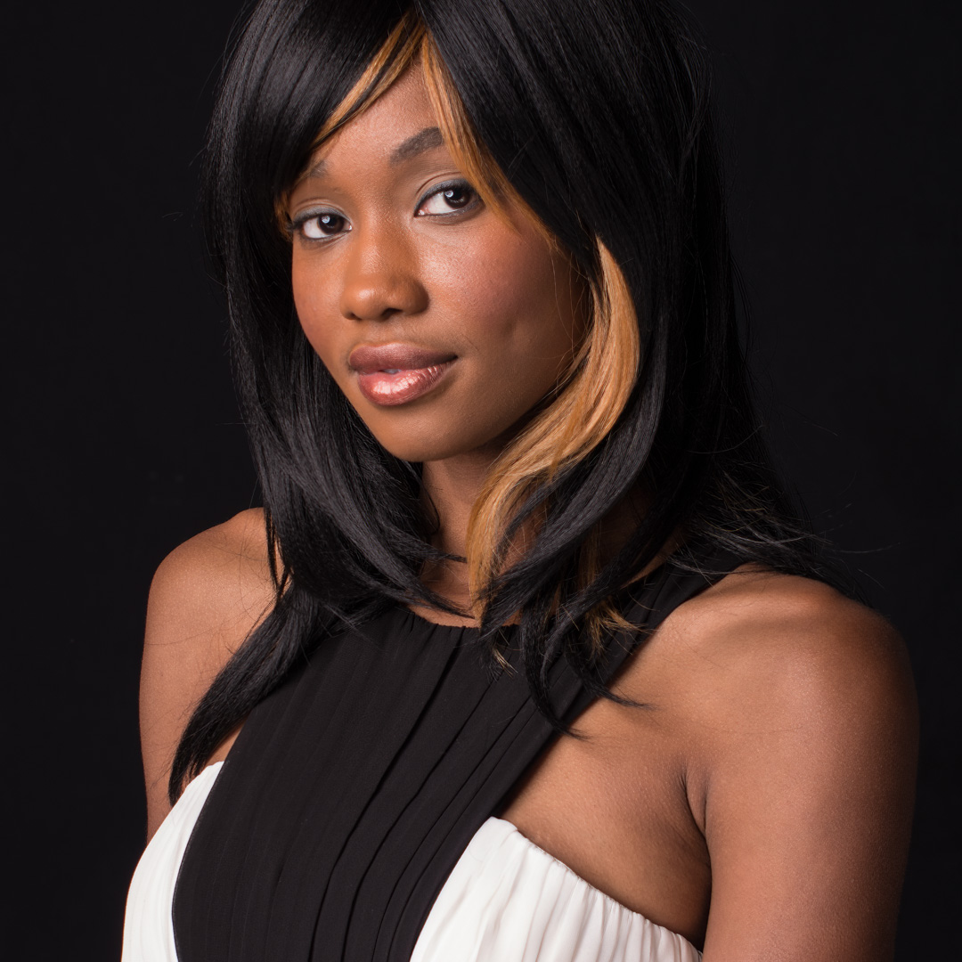 A woman with long black hair and blonde highlights poses against a black background. She wears a sleeveless black and white dress.