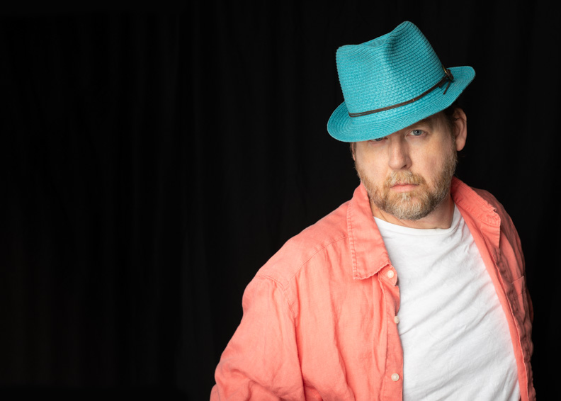 Person wearing a turquoise hat, coral shirt, and white t-shirt poses against a black background.